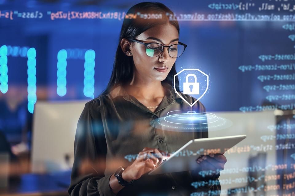 A young woman, likely a cybersecurity professional, concentrates on a tablet, while a digital holographic projection of a padlock emphasizes data security.